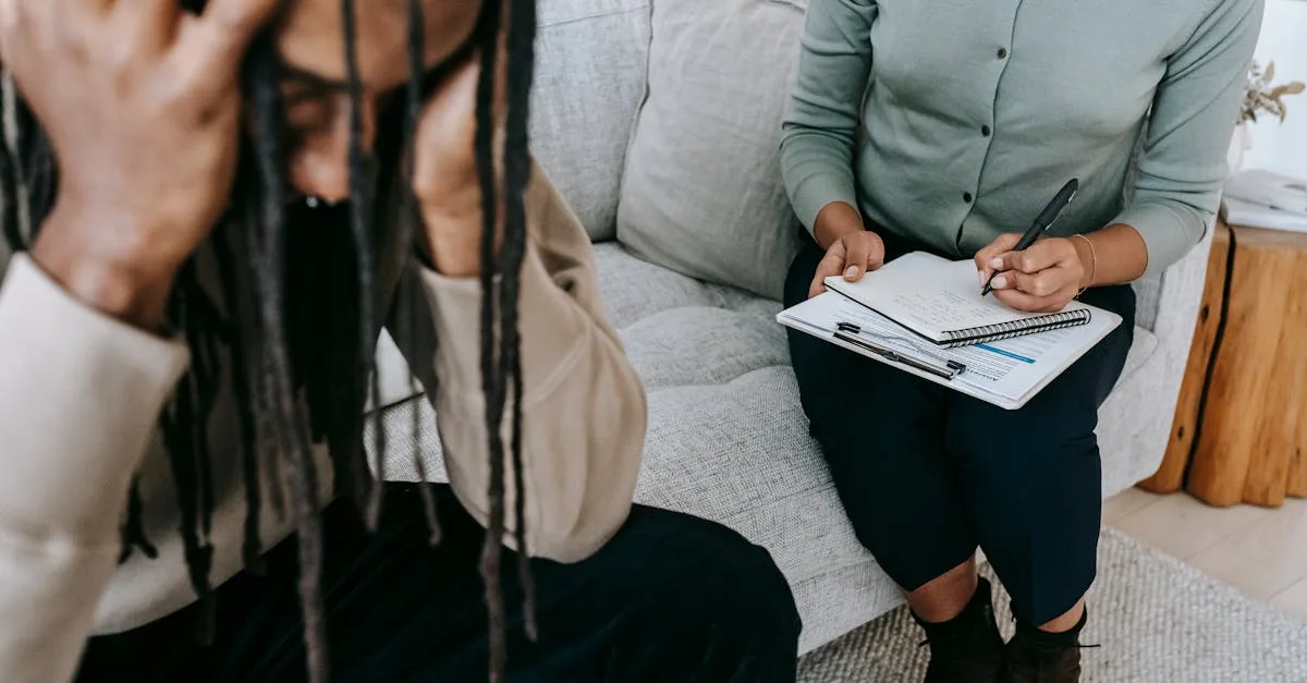 One-on-one therapy session: client sits on a couch with hands on head, while the therapist writes notes in a notebook on her lap.