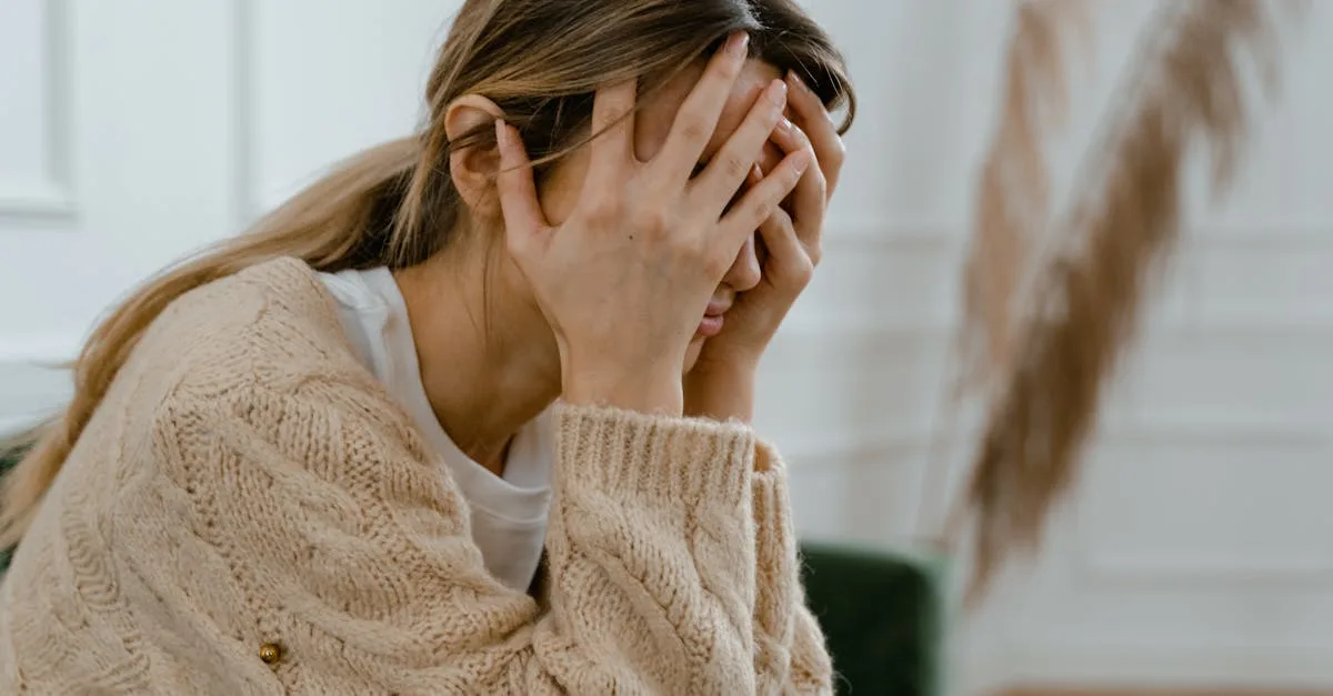 Woman in a beige knit sweater sits with her hands covering her face, appearing stressed or with anxiety.