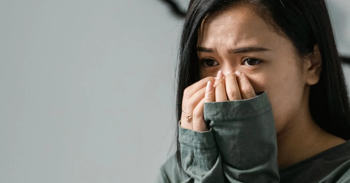 Young woman with long dark hair covers her mouth with her hands, appearing worried or distressed.