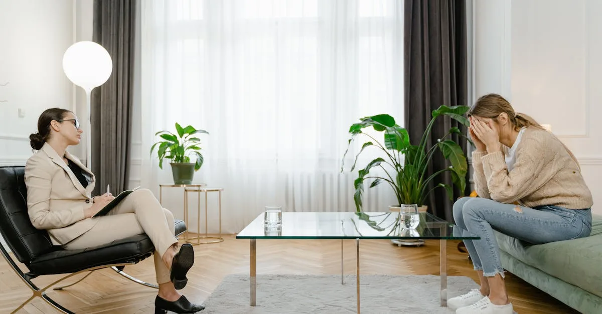 Therapist in a beige suit sits with a clipboard across from a distressed client covering her face in a bright living room with a glass coffee table and plants.