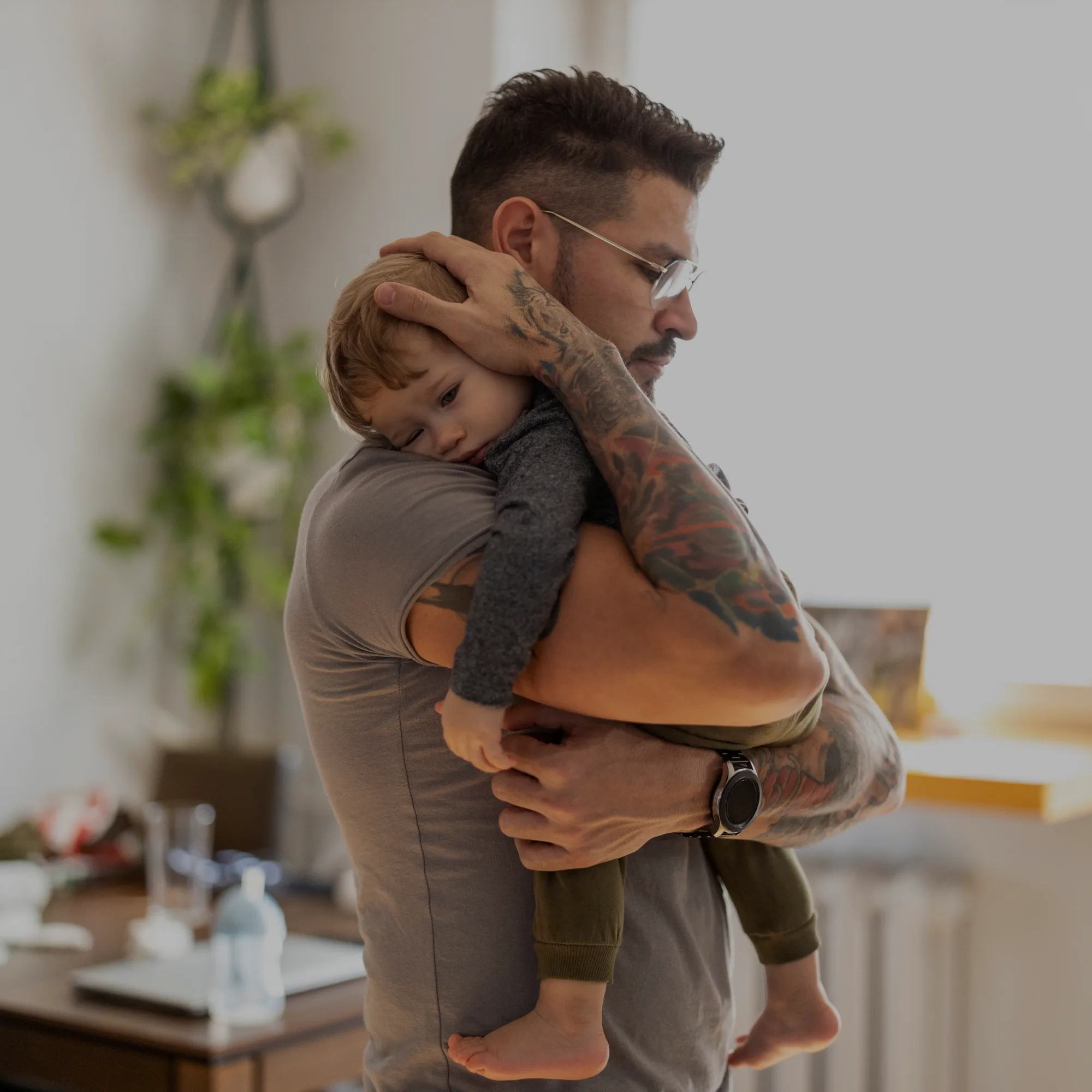 Man with tattoos hugging a small child close to his chest indoors, both facing left.