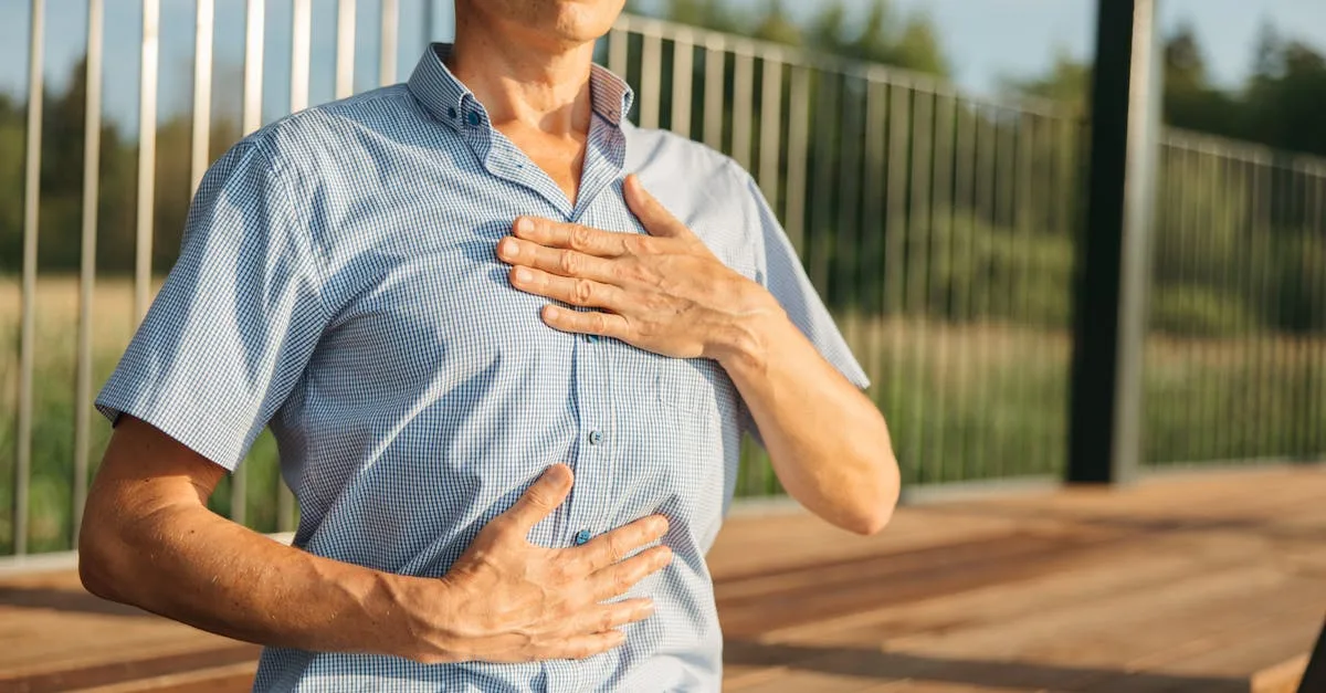 Man in a blue checkered shirt places a hand on his chest outdoors, suggesting chest pain or discomfort.