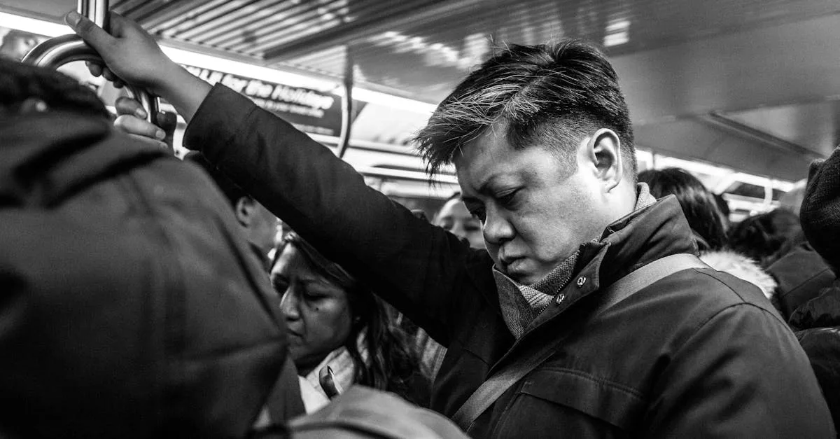 Man in a coat grips a metal handrail on a crowded subway car, looking downward.