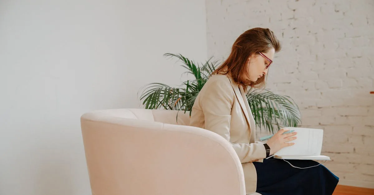 Woman in a beige blazer sits on a pale pink chair and reads a book in a bright, minimalist room with a plant nearby.