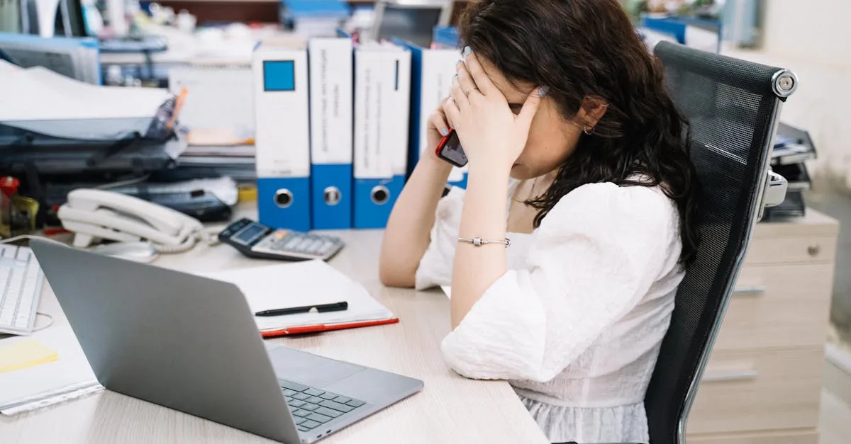 Woman at a cluttered office desk, head in hands while talking on a mobile phone beside a laptop and binders in the background.