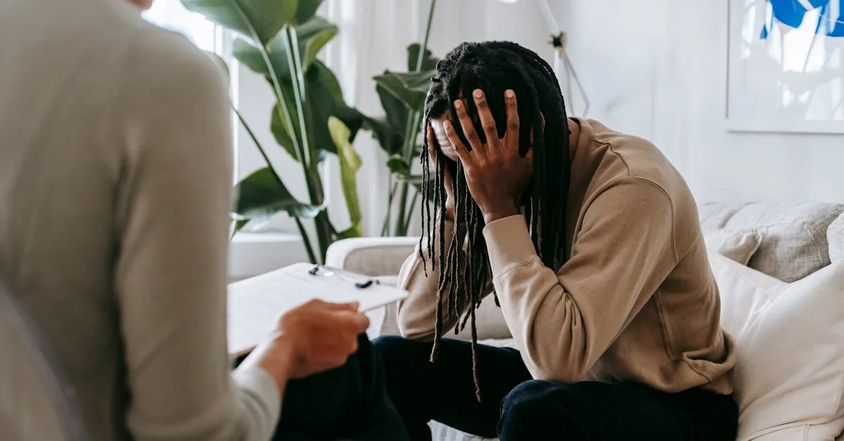 Person on a couch holds their head in their hands during a therapy session; a clinician with a clipboard sits nearby.