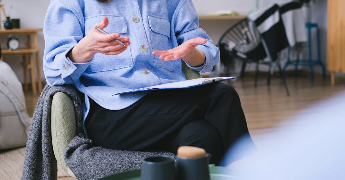 Person wearing a blue shirt gestures with hands while seated, with a clipboard on their lap in a cozy room.