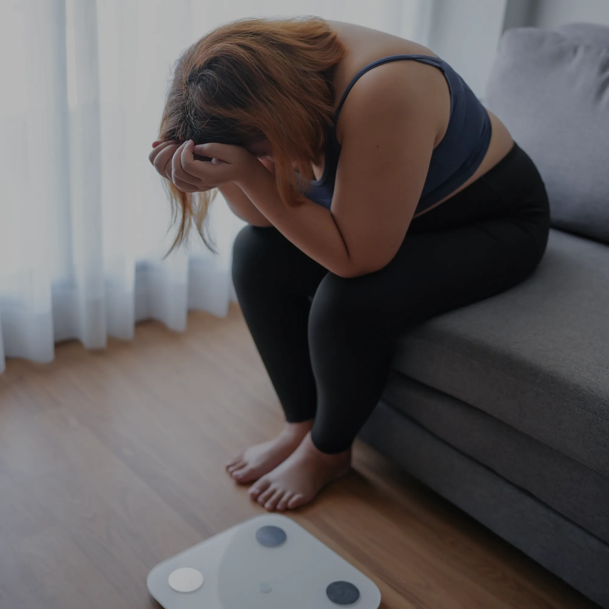 Person sitting on a couch with head in hands, looking distressed or overwhelmed, near a scale on the floor.