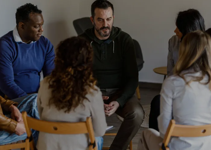 Five adults seated in a circle, engaged in a thoughtful discussion in a softly lit room.