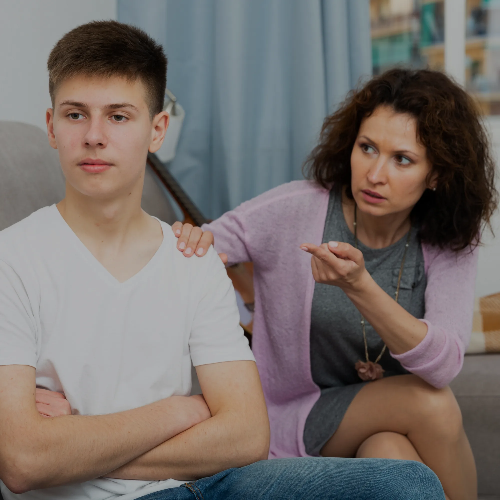 Two people sit together in a cozy room: a young man on the left crosses his arms while a woman to his right rests a hand on his shoulder and speaks softly.