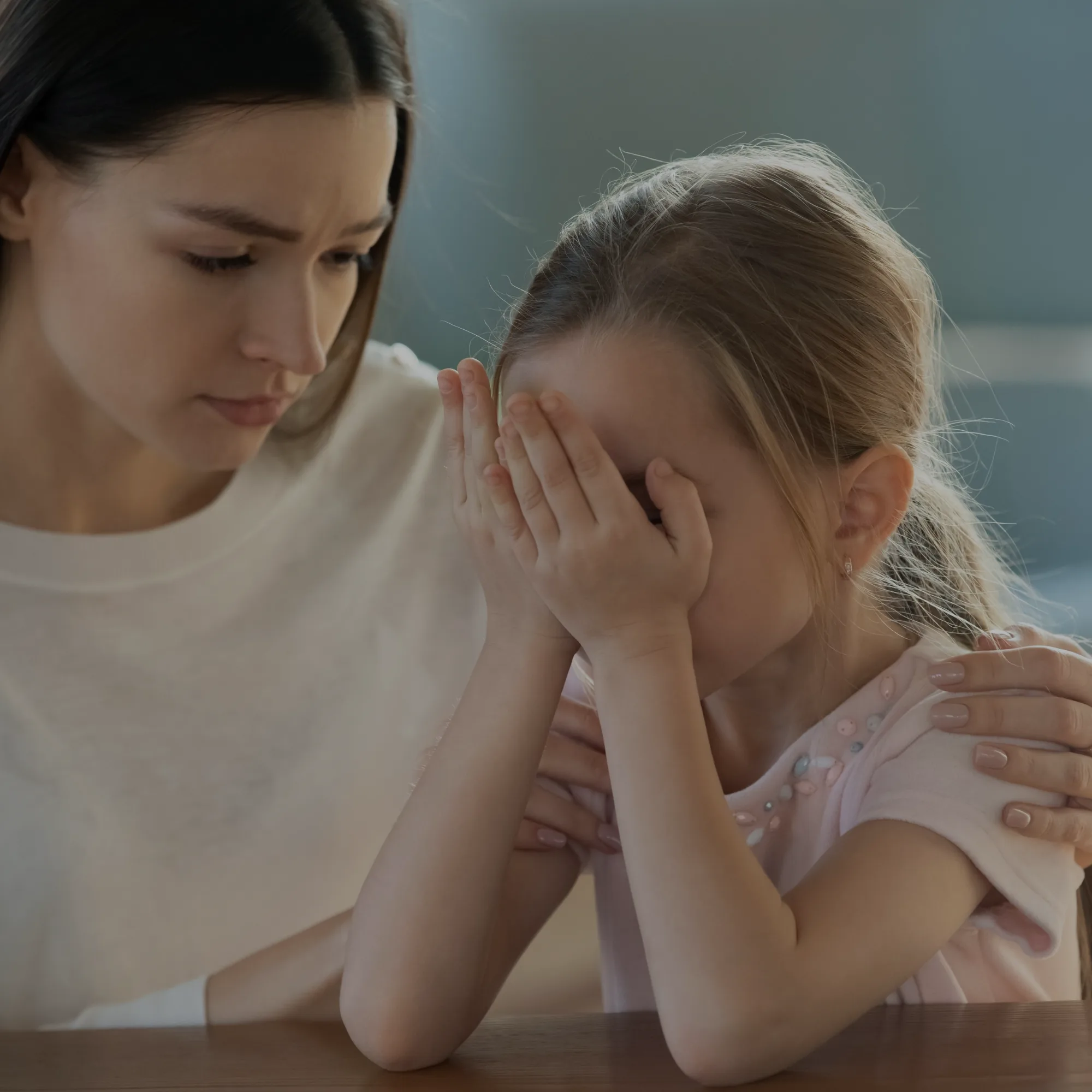 Adult and child share a comforting moment at a table; the child covers their face with hands while the adult offers support.