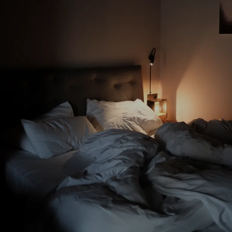 Dimly lit bedroom with an unmade bed, a padded headboard, and a bedside table with a warm lamp to the right.