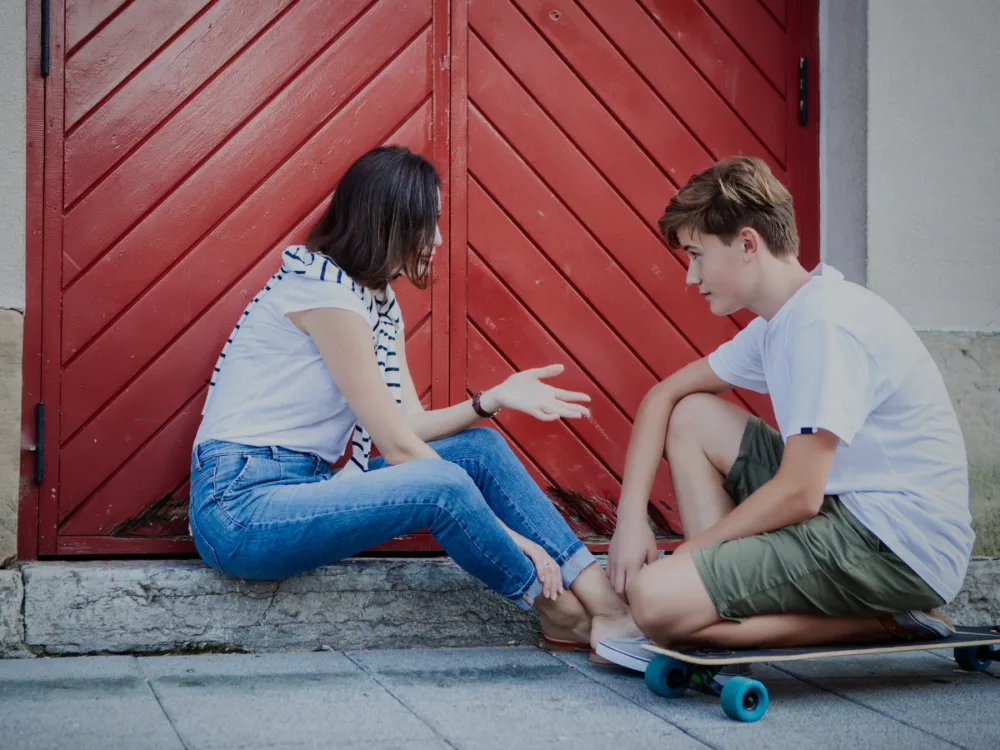 Two teens sit on the curb talking by a red door; the girl extends a hand toward the boy, skateboard nearby.