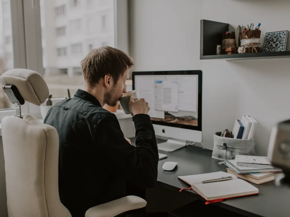 A person in front of a workspace, drinking a cup of coffee.