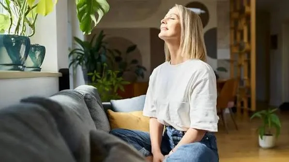 Woman relaxing on a gray sofa with eyes closed, basking in sunlight in a bright living room with plants nearby.
